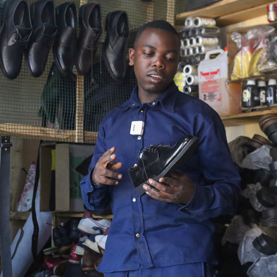 Ivan in his shoe workshop in Rwanda, holding a handmade black shoe, surrounded by leather materials and finished footwear.