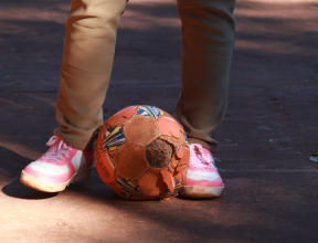 A worn soccer ball sits on the ground in front of a child wearing pink sneakers and beige pants.