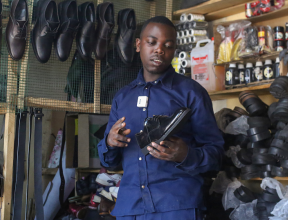 Ivan in his shoe workshop in Rwanda, holding a handmade black shoe, surrounded by leather materials and finished footwear.