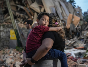 Mother holding child tightly in front of rubble caused by earthquake