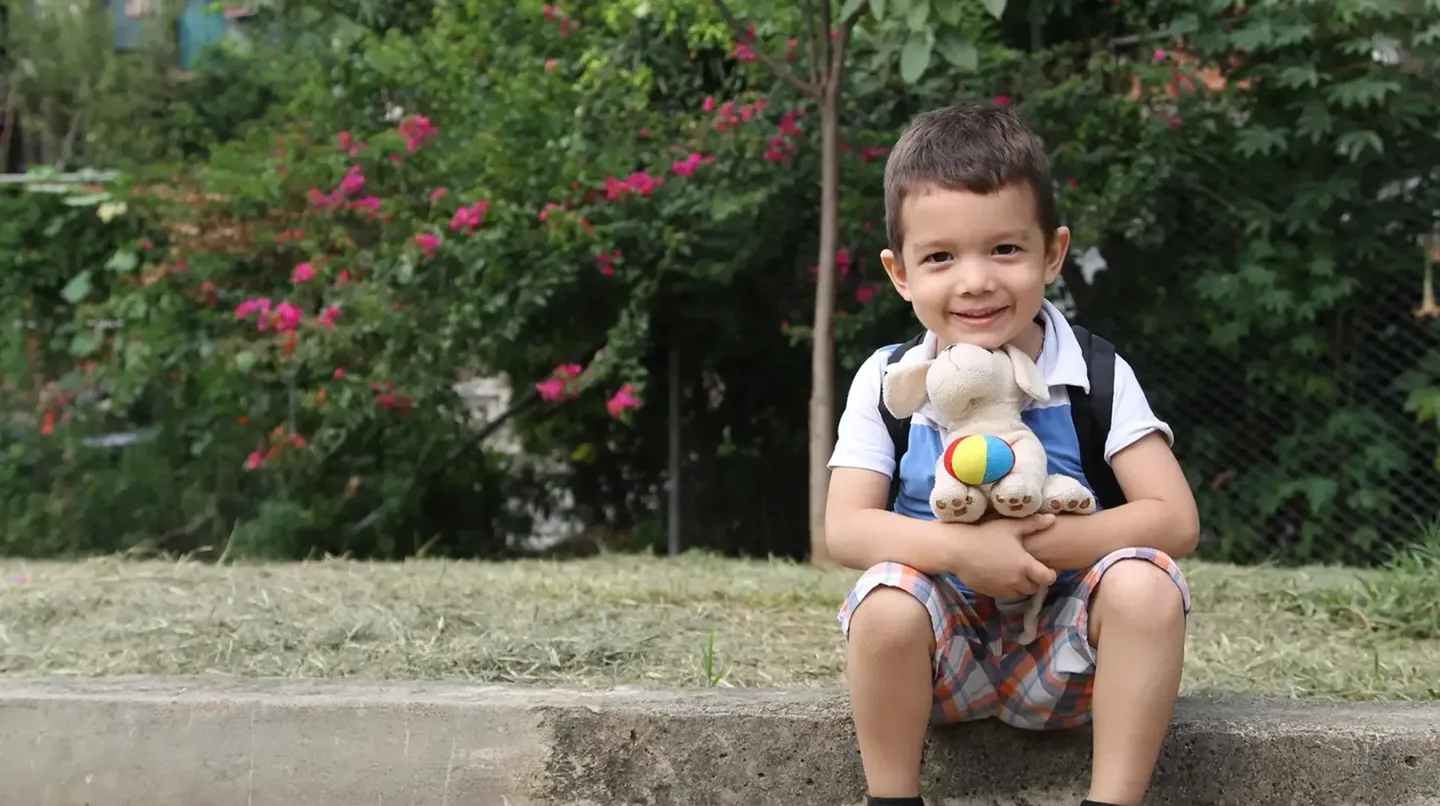 A boy sitting on a curb wearing a backpack and holding a stuffed animal