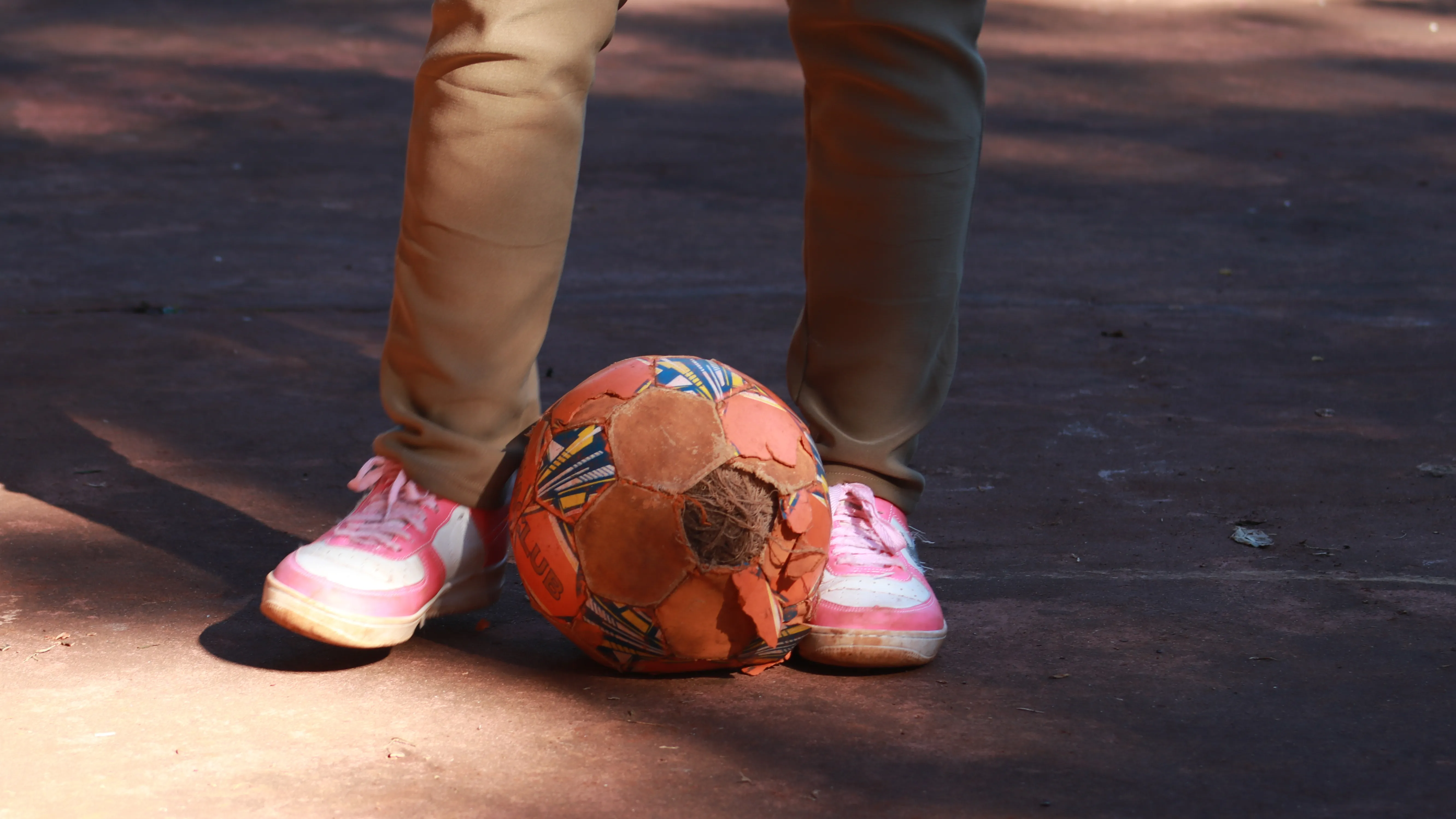 A worn soccer ball sits on the ground in front of a child wearing pink sneakers and beige pants.