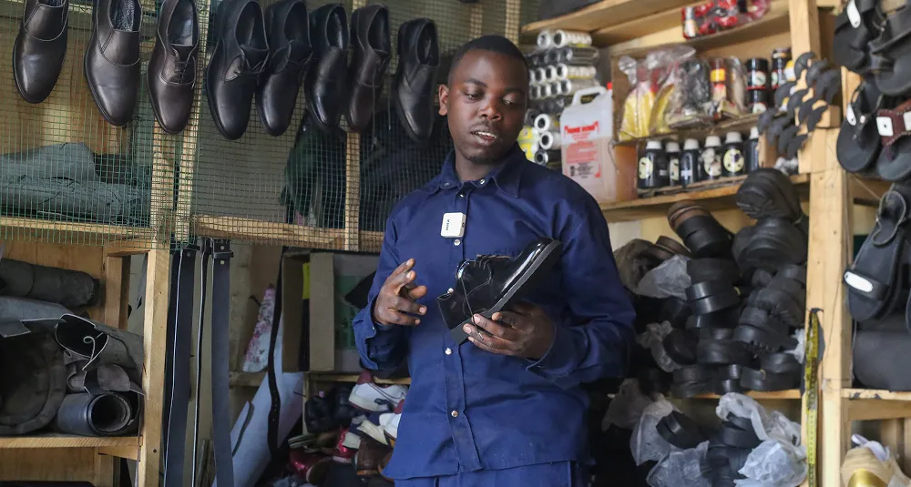 Ivan in his shoe workshop in Rwanda, holding a handmade black shoe, surrounded by leather materials and finished footwear.