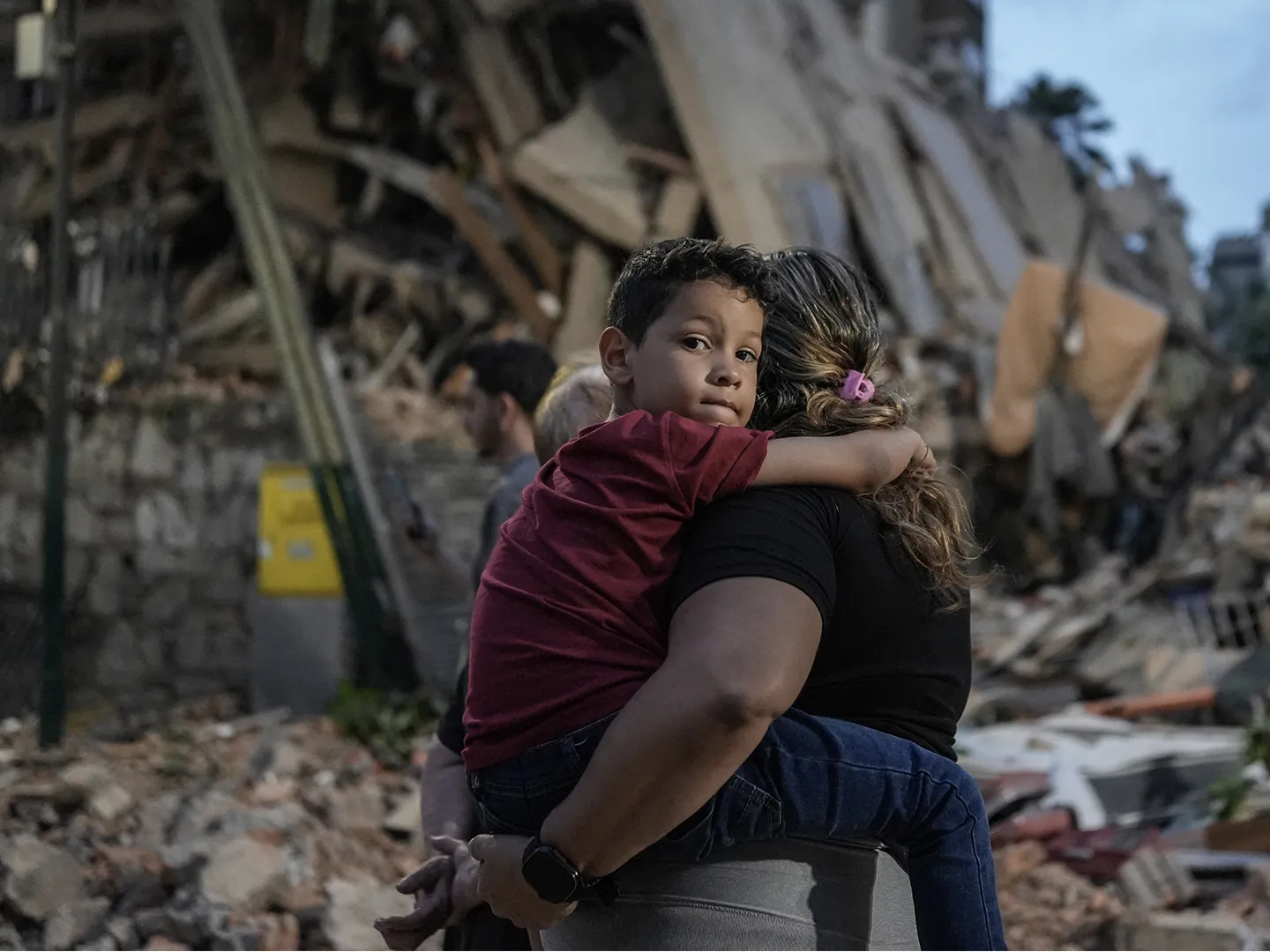Mother holding child tightly in front of rubble caused by earthquake.