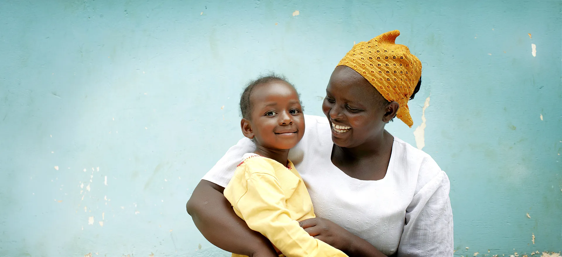 A smiling mother holding her young child against a light blue wall.  Photo credit: Jens Honoré.